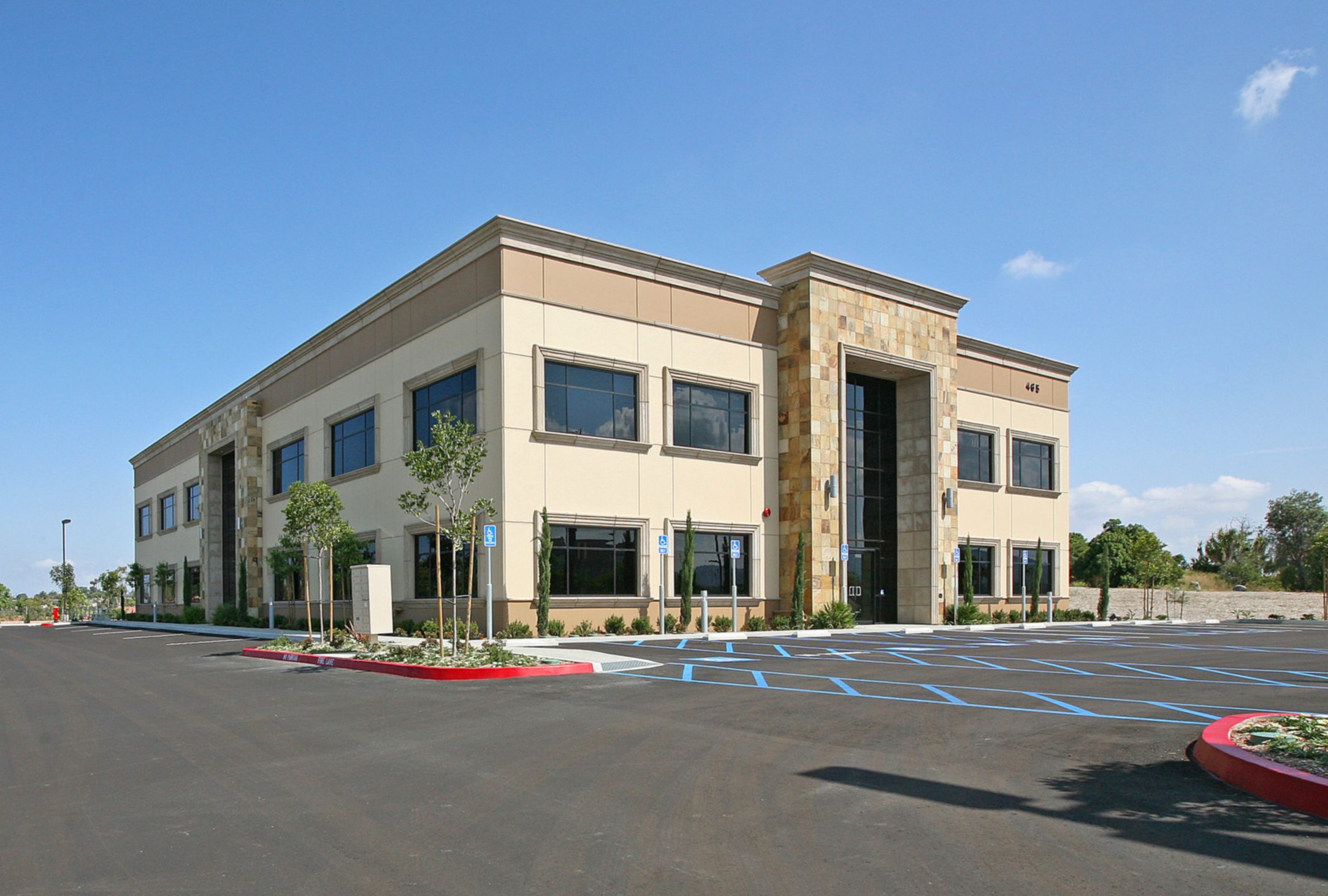 A two-story commercial building with large windows, beige exterior, and a central entrance, surrounded by an empty parking area under a clear blue sky.