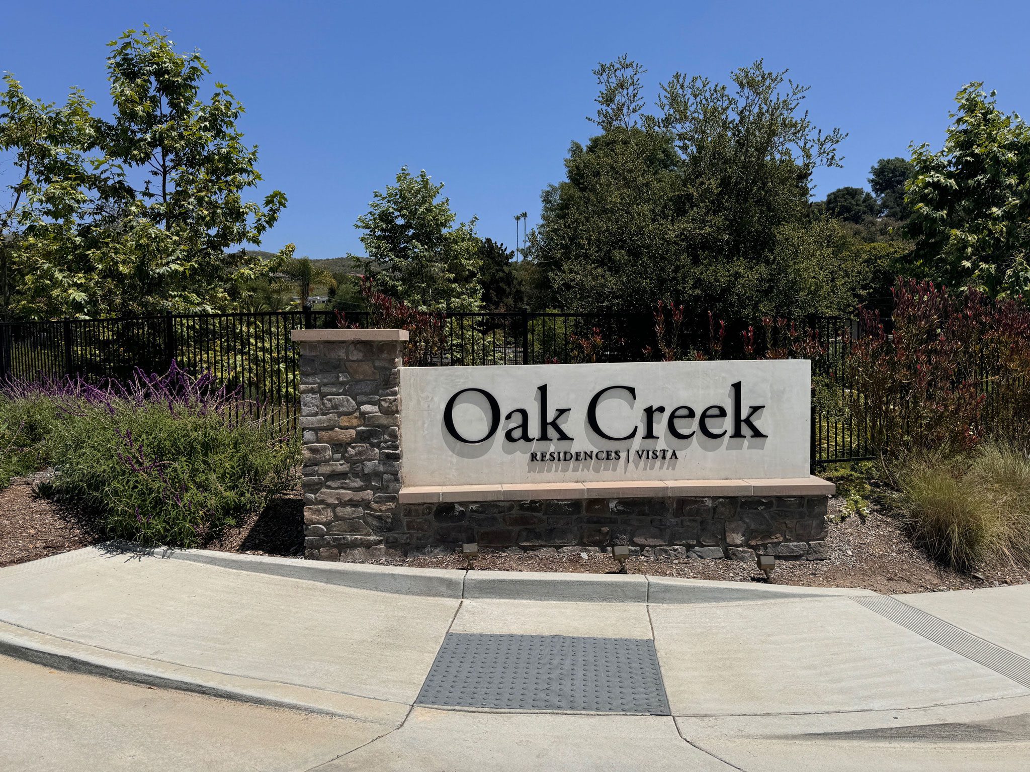 A sign with "Oak Creek Residences Vista" inscribed on it, situated next to a stone pillar and surrounded by greenery and a black fence under a clear blue sky.