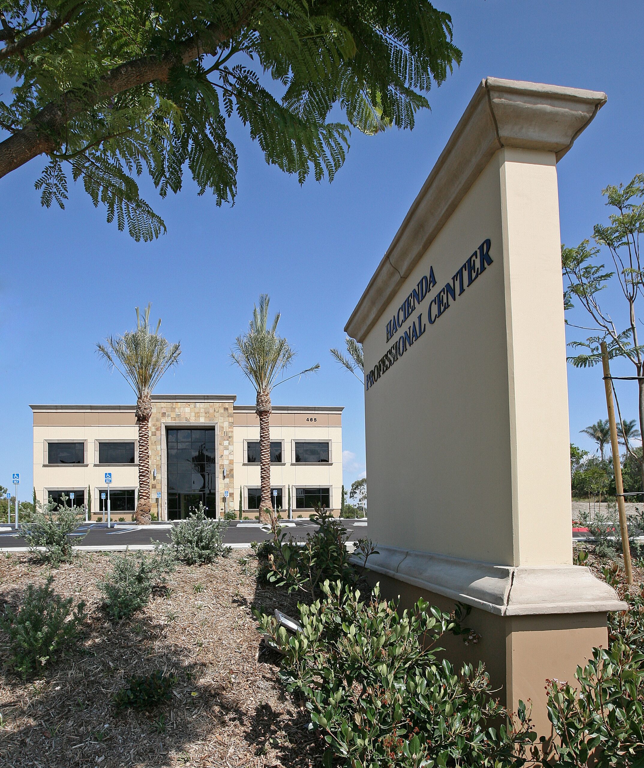 A beige sign reading "Hacienda Professional Center" stands in front of a two-story office building with palm trees and landscaped grounds under a clear blue sky.