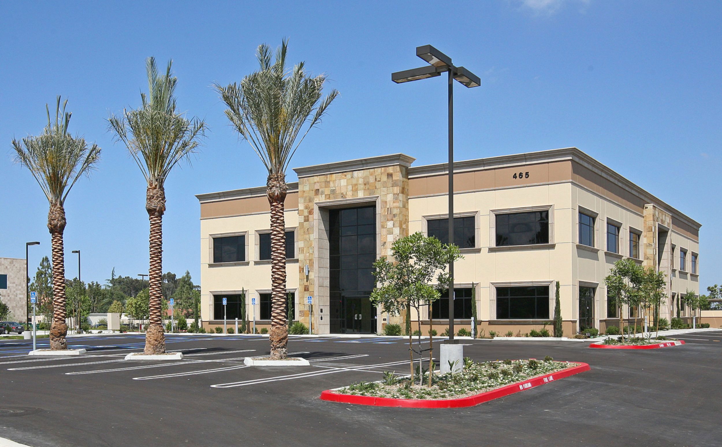 A two-story commercial building with large windows and stone accents, surrounded by an empty parking lot with palm trees and small plants. The building number "465" is visible.