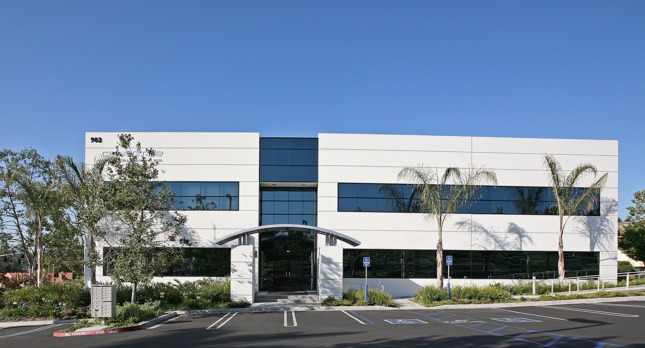 Modern two-story office building with large reflective windows and an arched entrance, surrounded by palm trees and a parking lot with marked parking spaces.