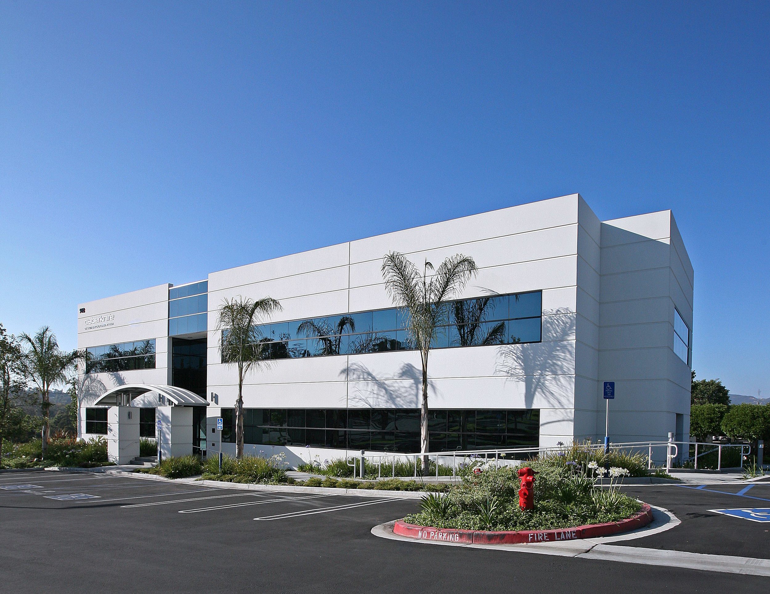 A modern two-story office building with large windows, palm trees in front, and a landscaped parking area under a clear blue sky.