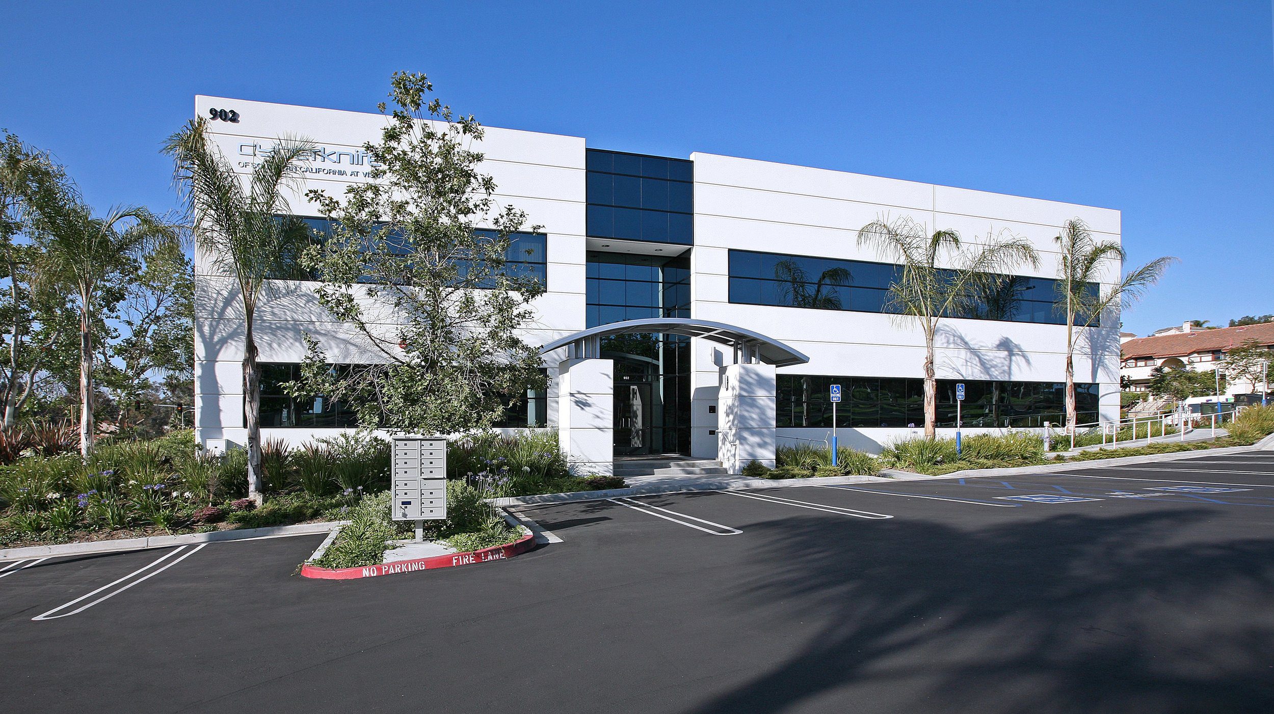 A modern, two-story commercial building with large windows and an arched entrance, surrounded by palm trees and landscaping, with an adjacent parking lot under a clear blue sky.